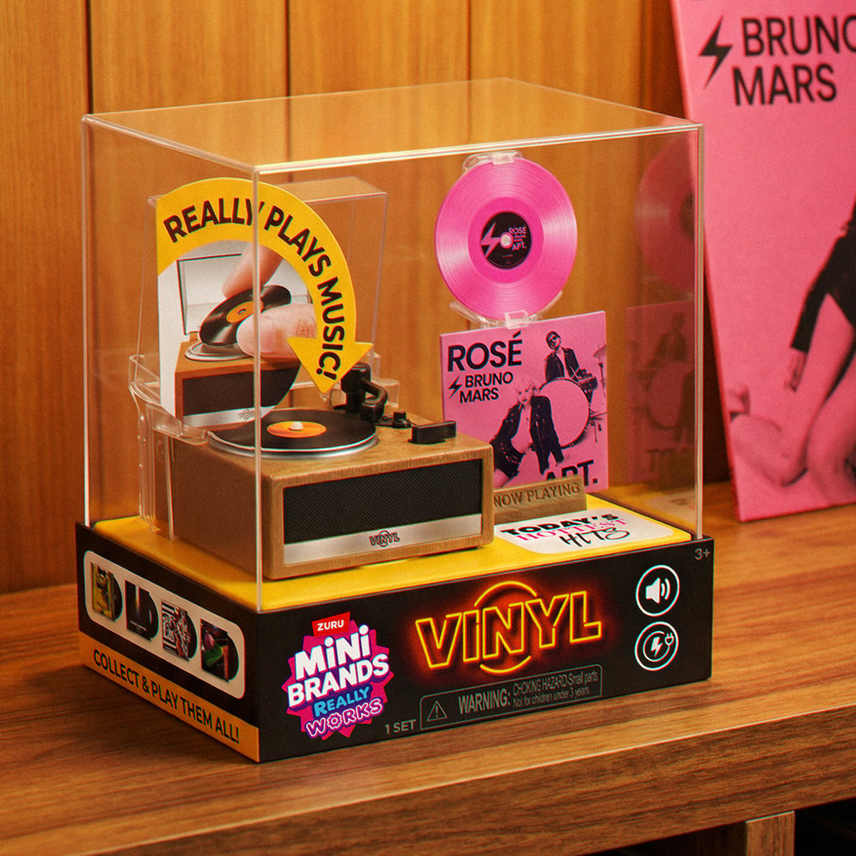 Verpacktes ZURU Mini Brands Vinyl-Spielset auf einer Holzfläche vor einer Backsteinwand.